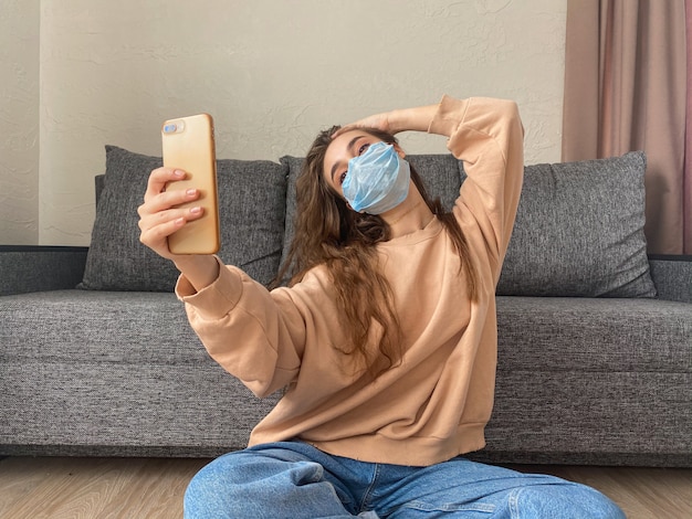 Chica atractiva con una máscara médica toma una selfie en un teléfono inteligente sentado en su casa en autoaislamiento debido a la pandemia de coronavirus