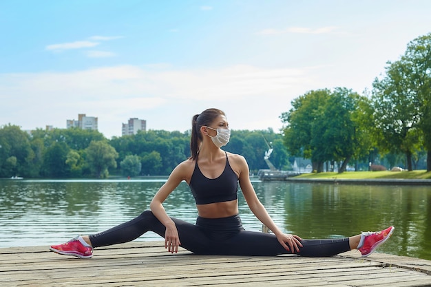 Chica atractiva con una máscara y ropa deportiva está haciendo ejercicios de gimnasia en un muelle de madera en un parque de la ciudad. concepto de entrenamiento al aire libre seguro