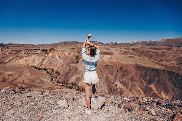 Una chica atractiva con pantalones cortos y una camisa se para con la espalda con las manos levantadas y apoyada en una piedra se encuentra a un lado en una vista rocosa del cañón bajo un cielo claro y sin nubes.