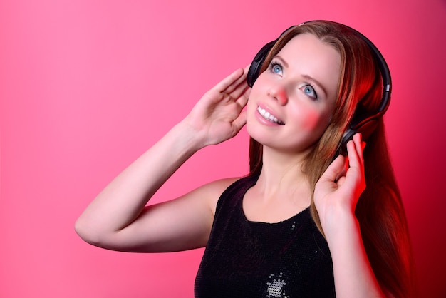 Foto chica atractiva con el pelo largo en grandes auriculares inalámbricos escuchando música sobre un fondo rosa