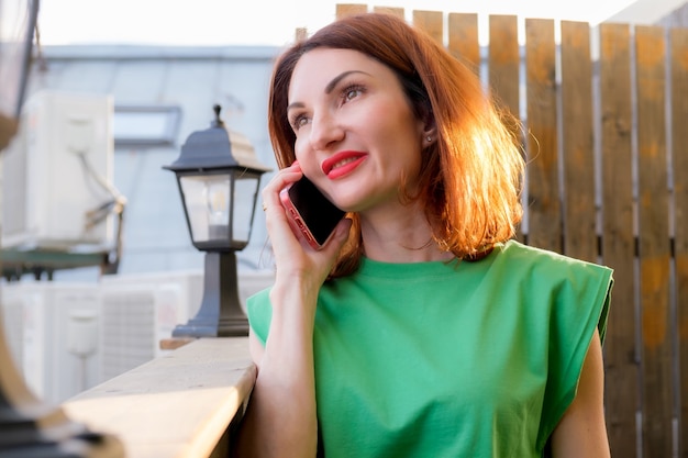 Chica atractiva con el pelo rojo en una blusa verde está hablando por un teléfono móvil en la terraza abierta de un café. La mujer de negocios está negociando por teléfono.