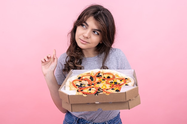 Chica atractiva con pizza en una caja para entrega sobre un fondo rosa.