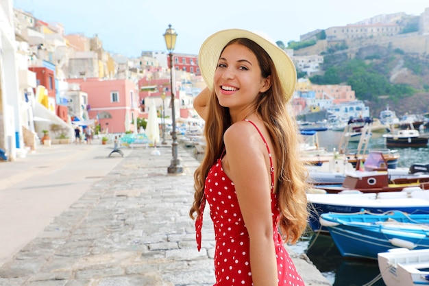 Chica atractiva en Procida, Italia. Hermosa mujer de moda con vestido rojo de lunares y sombrero camina por el puerto de la isla de Procida, capital italiana de la cultura 2022.