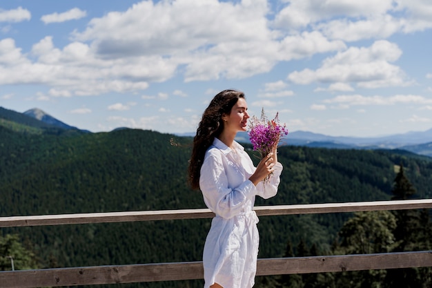 Chica atractiva con ramo de flores en las colinas verdes Mujer joven weared vestido blanco sosteniendo flores rosadas
