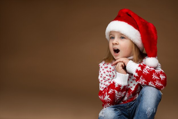 Chica atractiva en ropa acogedora y sombrero de Navidad se sienta en el suelo y canta una canción de Navidad, imagen aislada sobre fondo marrón