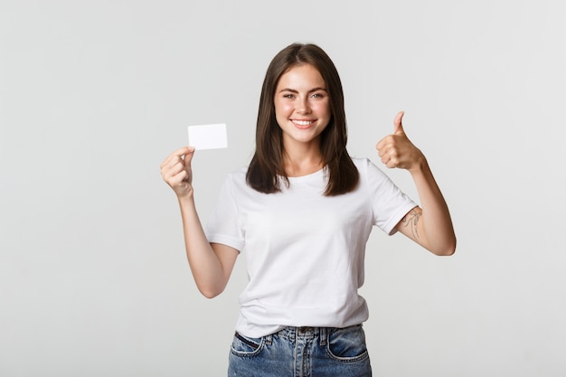 Chica atractiva satisfecha mostrando tarjeta de crédito y pulgar hacia arriba