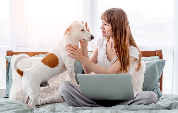 Chica atractiva sentada en la cama con portátil gris y acariciando a su lindo perro. Mujer hermosa joven con mascota y portátil en la mañana en el dormitorio. Concepto de trabajo remoto desde casa