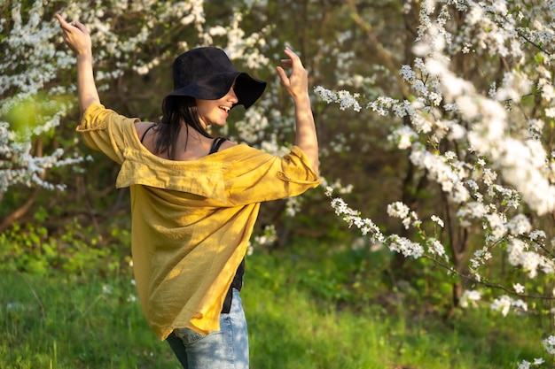 Una chica atractiva con un sombrero entre árboles en flor disfruta del olor de las flores de primavera.