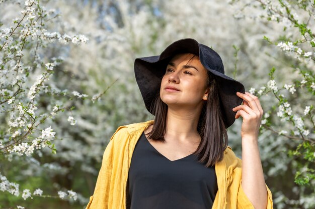 Chica atractiva con un sombrero entre los árboles en flor en la primavera, en un estilo casual