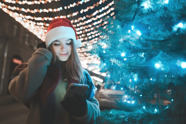 Chica atractiva con un sombrero de Navidad se encuentra en el árbol de Navidad de la calle, smartphone en sus manos, mirando la pantalla del smartphone, fondo de adornos navideños de la calle