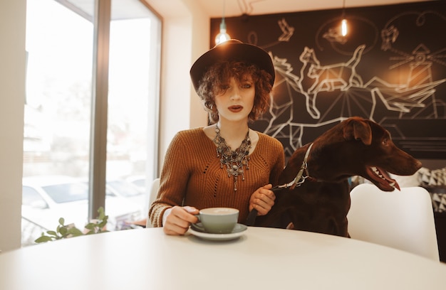 Foto chica atractiva con un sombrero y con el pelo corto y rizado se sienta en un acogedor café con un perro