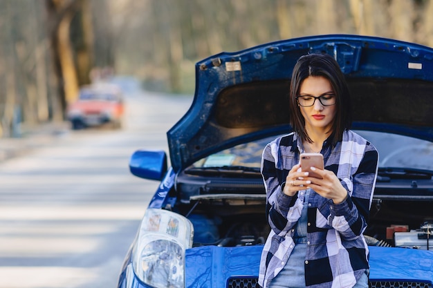 Chica atractiva con teléfono cerca de capó abierto de coche