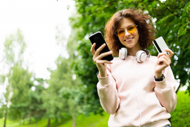 Chica atractiva con un teléfono y una tarjeta de crédito en una pared del parque verde