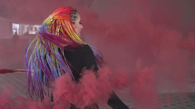 Foto chica atractiva con trenzas multicolores y maquillaje en un vestido negro. posando escondido detrás de humo artificial rojo contra el fondo de un edificio en una ciudad primaveral.