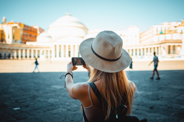 Foto chica atractiva turista con sombrero con mochila explorando la nueva ciudad de europa en verano y usando su teléfono para tomar fotos