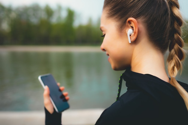 Chica atractiva usando su teléfono, auriculares inalámbricos mientras se ejecuta en el parque