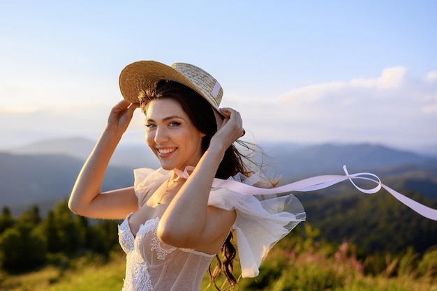 Chica atractiva en vestido de novia y sombrero mirando a la cámara en las montañas