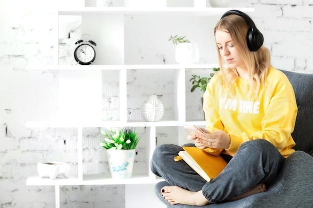Chica en auriculares en blusa amarilla sentarse, relajarse en el sofá gris, usar el teléfono inteligente y la nota para escuchar música