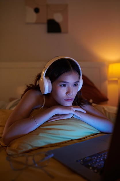 Chica con auriculares viendo melodrama en una computadora portátil mientras está acostada en la cama