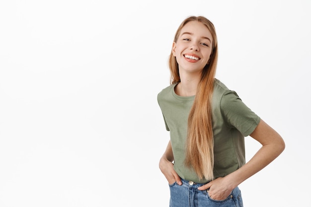 Chica auténtica despreocupada con sonrisa blanca perfecta emociones felices tomados de la mano en jeans y mirando alegre a la cámara de pie contra el fondo blanco