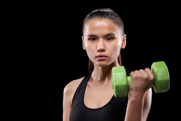 Chica bastante activa sosteniendo mancuernas verdes en la mano mientras hace ejercicio en el gimnasio o centro deportivo sobre pared negra