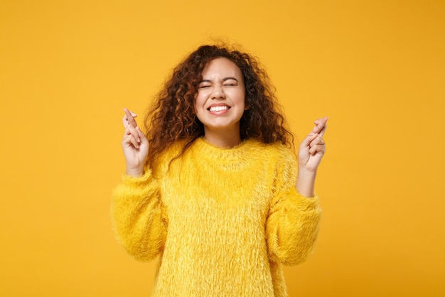 Chica bastante afroamericana posando aislada sobre fondo amarillo naranja. Concepto de estilo de vida de las personas. Simulacros de espacio de copia. Esperando un momento especial, manteniendo los dedos cruzados, los ojos cerrados, pidiendo un deseo.