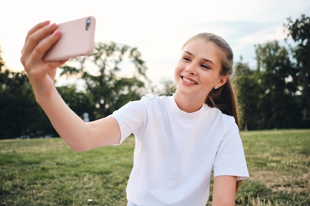 Chica bastante alegre con camiseta blanca felizmente tomando fotos en el celular mientras se sienta en el césped en el parque de la ciudad