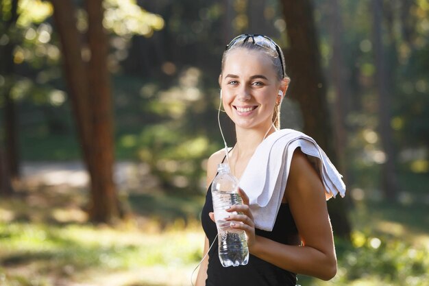 Chica bastante atlética con toalla escuchando música en el parque después de correr y sostener una botella con agua buen día soleado