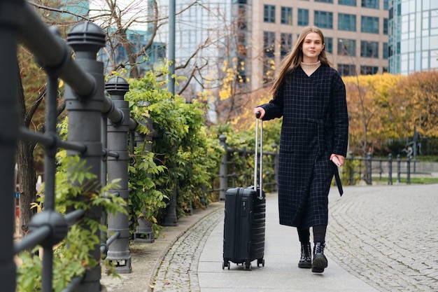 Chica bastante elegante en abrigo caminando soñadoramente por la calle de la ciudad con maleta