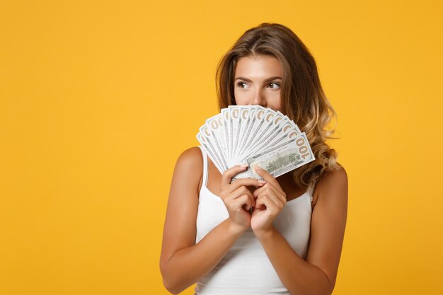 Chica bastante joven con ropa informal ligera posando aislada en el fondo de la pared naranja amarilla en el estudio. Concepto de estilo de vida de las personas. Simulacros de espacio de copia. Sosteniendo fan de dinero en efectivo en billetes de dólar.