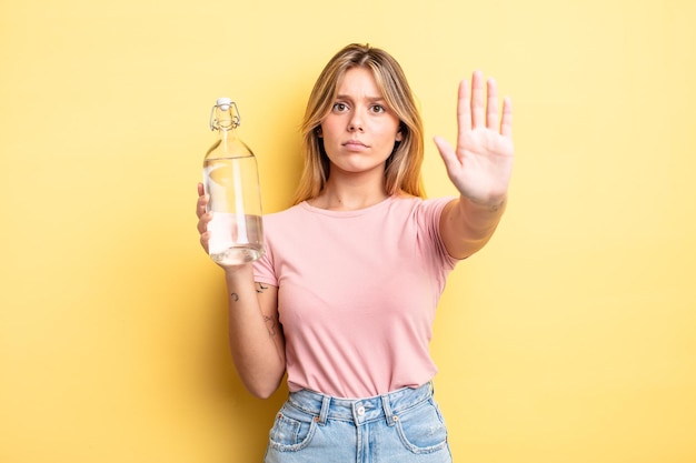 Chica bastante rubia mirando seria mostrando la palma abierta haciendo gesto de parada. concepto de botella de agua