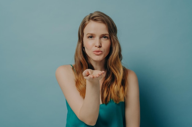 Una chica bastante sincera que sopla un beso de aire y expresa amor mientras está aislada de fondo azul