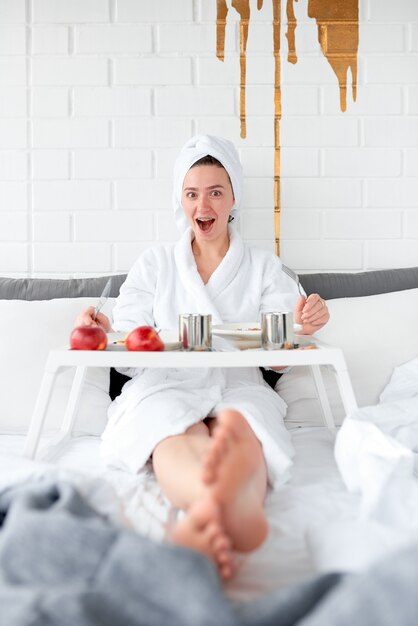 La chica en bata de baño tiene un tenedor y un cuchillo en sus manos y está lista para un delicioso desayuno en la cama