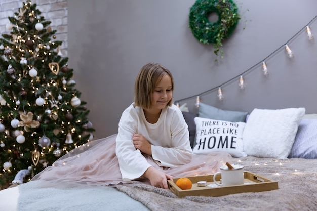 Chica bebe té y come en la cama en el dormitorio en medio del árbol de Navidad festivo, enfoque suave