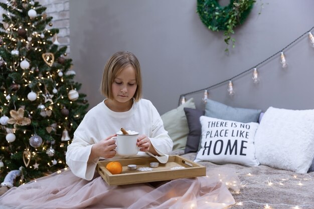 Chica bebe té y come en la cama en el dormitorio en medio del árbol de Navidad festivo, enfoque suave