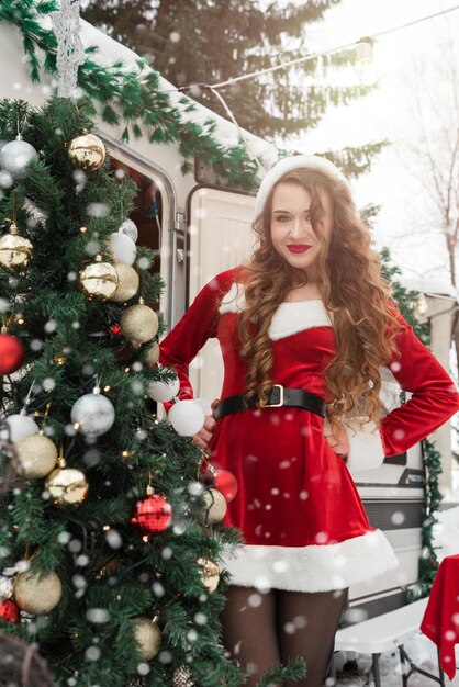 Chica de belleza en traje de santa decora el árbol de navidad en el campamento de invierno preparándose para la n