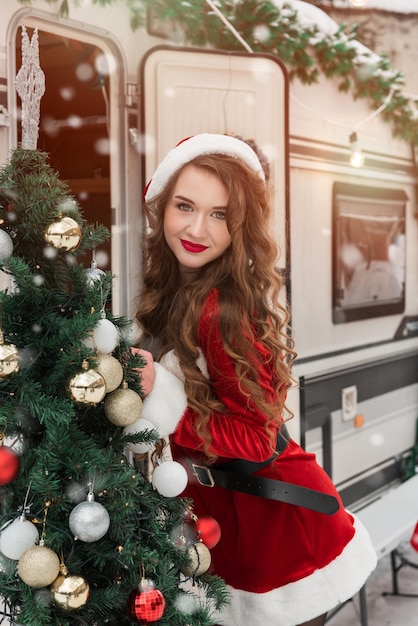 Chica de belleza en traje de santa decora el árbol de navidad en el campamento de invierno preparándose para la n