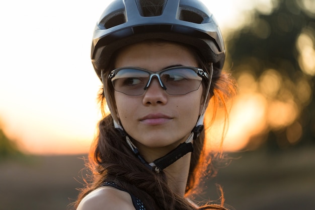 Chica en bicicleta de montaña en campo a través, hermoso retrato de un ciclista al atardecer, Chica fitness monta una bicicleta de montaña de fibra de carbono moderna en ropa deportiva. Primer plano, retrato, de, un, niña, en, un, casco, y, anteojos