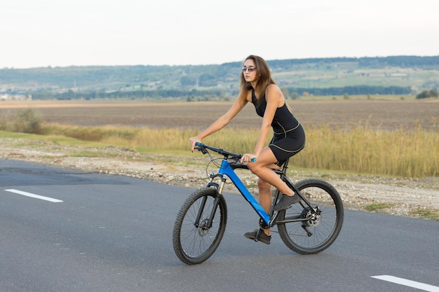 Chica en bicicleta de montaña en campo a través hermoso retrato de un ciclista al atardecer Chica fitness monta una bicicleta de montaña moderna de fibra de carbono en ropa deportiva Retrato de una niña con casco y gafas