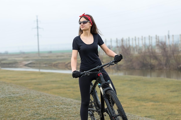 Chica en bicicleta de montaña en campo a través hermoso retrato de un ciclista en clima lluvioso Chica fitness monta una bicicleta de montaña moderna de fibra de carbono en ropa deportiva Retrato de una niña en pañuelo rojo