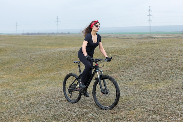 Chica en bicicleta de montaña en campo a través hermoso retrato de un ciclista en clima lluvioso Chica fitness monta una bicicleta de montaña moderna de fibra de carbono en ropa deportiva Retrato de una niña en pañuelo rojo