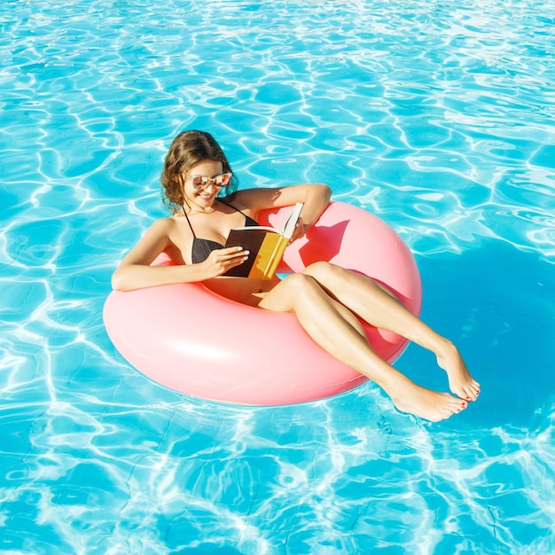 Chica en bikini con gafas de sol relajada y leyendo un libro sobre un anillo de piscina inflable rosa