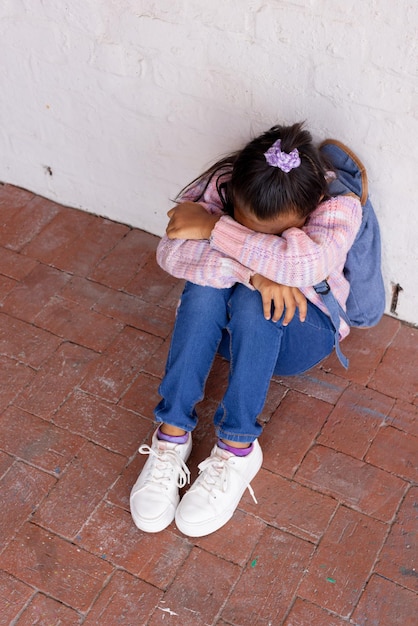 Una chica biracial se sienta acurrucada en el suelo mirando molesta en la escuela