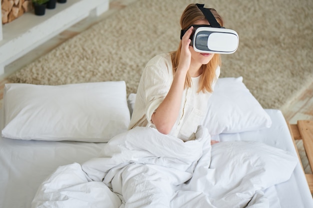 La chica de blanco sentada en la cama en el casco de realidad virtual