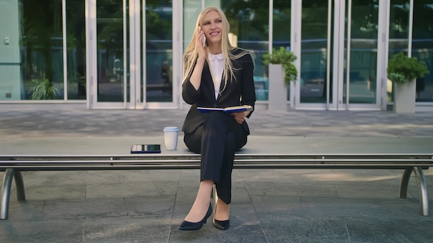 Chica con bloc de notas hablando por teléfono al aire libre. Mujer de negocios moderna en traje sentado en el banco con el bloc de notas en las manos y con llamada telefónica mirando a otro lado.