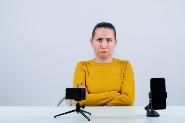 Chica blogger está mostrando mimetismo llorando mirando hacia arriba frente a su mini cámara sobre fondo blanco.