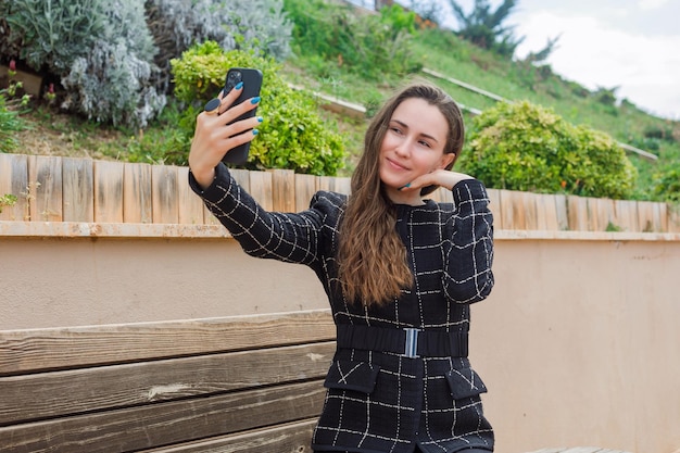 Chica bloguera se está tomando selfie con su teléfono inteligente poniendo la mano en la mejilla en el parque