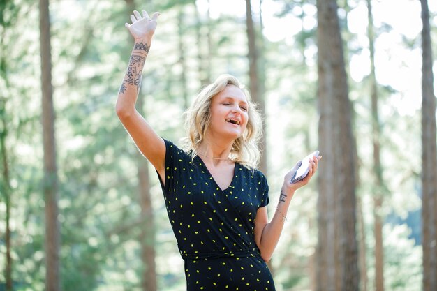 Chica bloguera feliz está bailando sosteniendo el móvil en el fondo de la naturaleza