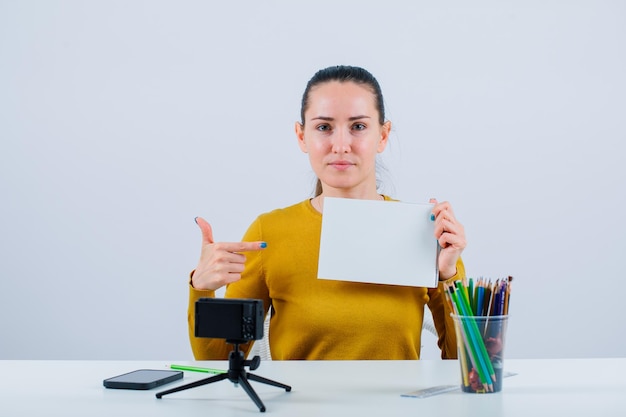 Chica bloguera sostiene papel blanco y lo muestra con el dedo índice sobre fondo blanco