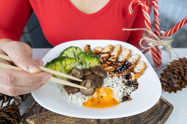 Una chica con una blusa roja y un delantal marrón sostiene un plato de shiitake con brócoli y pollo con arroz dorado